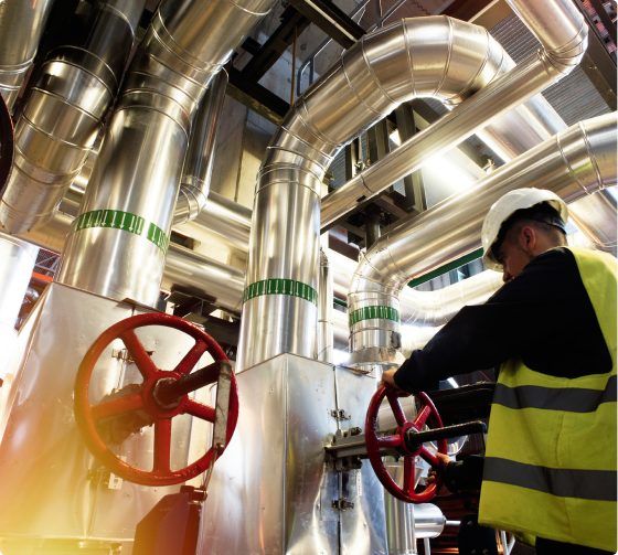 A worker wearing a hard hat and safety vest adjusts a red valve on a large network of industrial pipes.