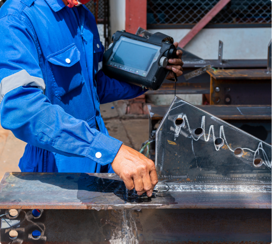 A person in a blue jumpsuit operates an ultrasonic testing device on a metal weld, checking for defects.